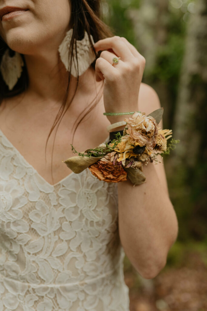 A bride is standing in the forest of North Carolina in her lacy elopement dress. She has on a floral wristlet and boho weaving earrings and a beautiful green ring.