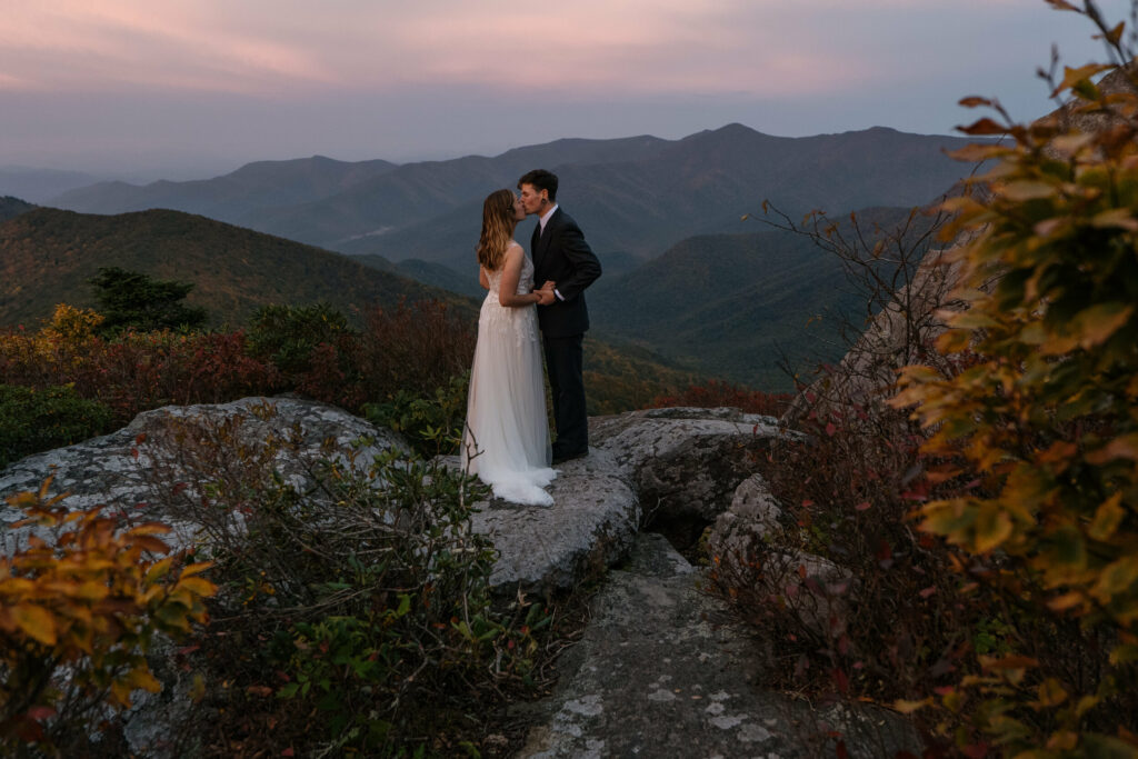 A couple is kissing in wedding clothes at sunrise on the Blue Ridge parkway, one of the best places to elope in Asheville NC. One is in a dress and the other in a black suit. There is fall foliage surrounding them and they are standing on a rock feature.