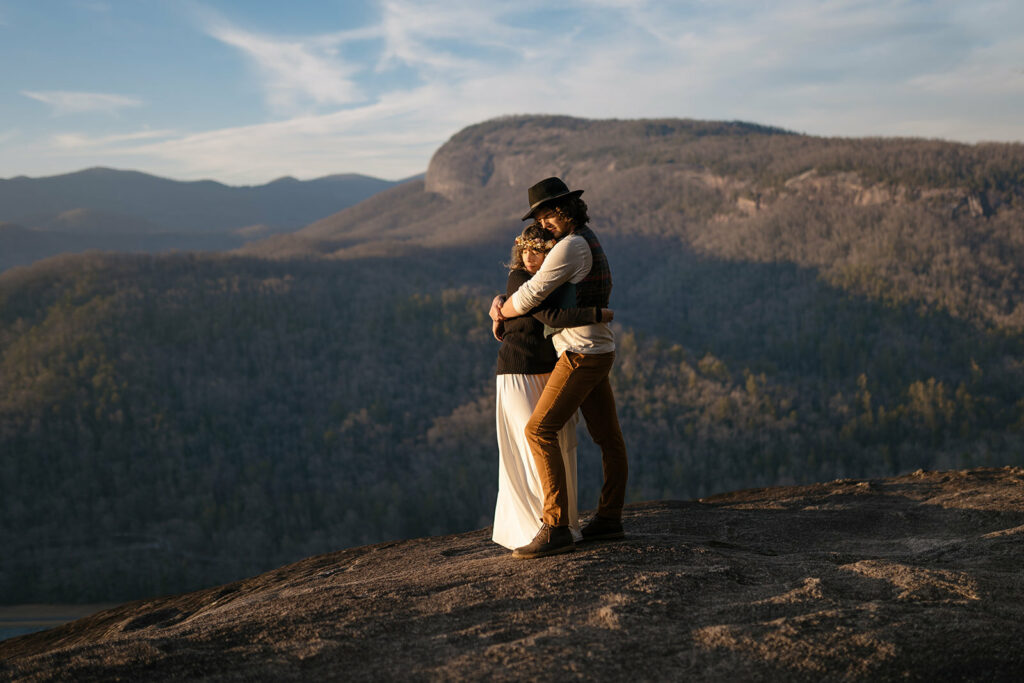 A couple stands in front of their Asheville elopement photographer and poses in a hug in front of john rock at sunset