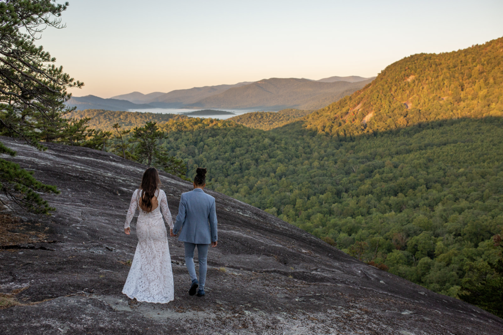 The photographer is standing behind the couple walking out on a rock ledge as the sunrise is lighting up the mountains in the background.