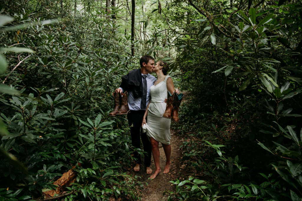 A shot while a couple walks through a rhododendron tunnel after taking a swim in the river in their wedding clothes. They kiss and are holding up their boots while they walk barefoot.