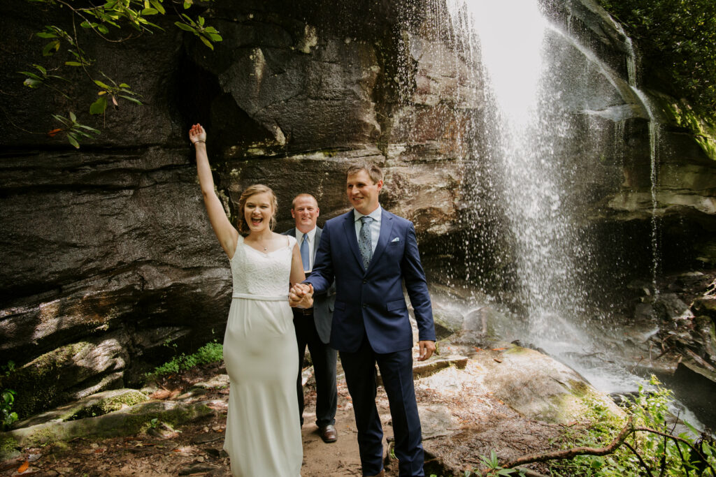 An Asheville elopement is captured by a photographer where the couple was just pronounced married in front of a waterfall. The bride has her arm raised in celebration.