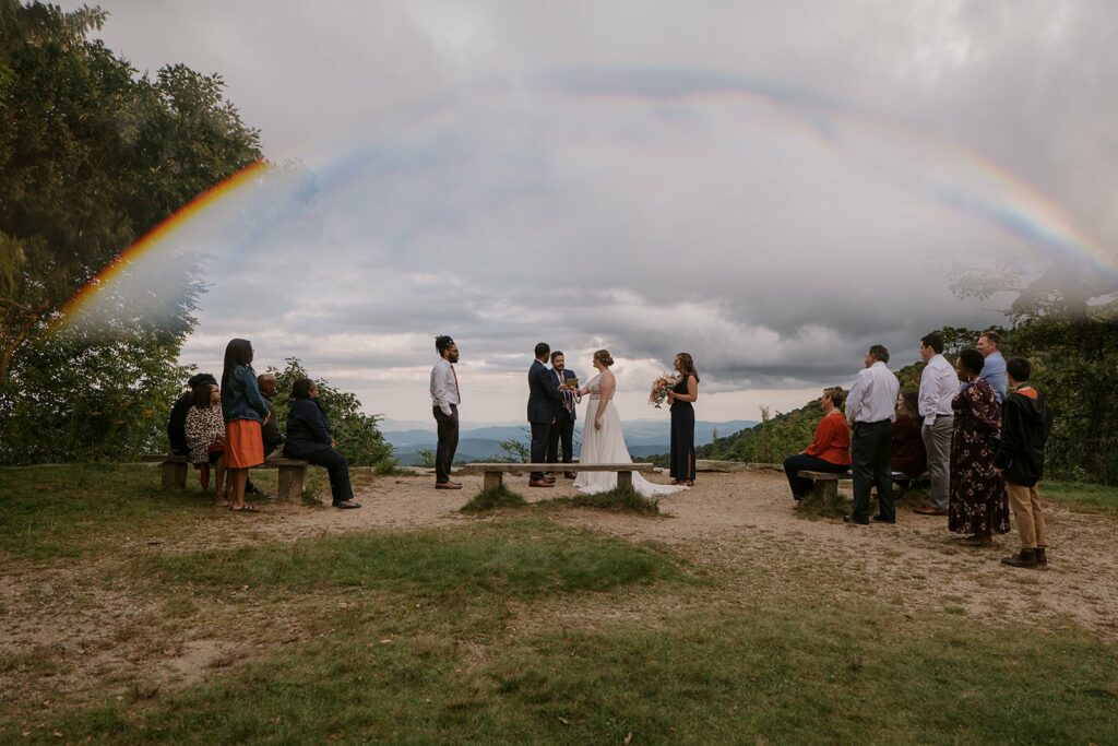 A rainbow sits above the heads of a family watching their loved ones have an elopement ceremony in the blue ridge mountains at an overlook with benches.