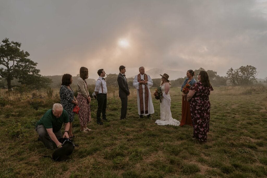 A small group of people are standing on a grassy hill with the sun shining through the fog during a ceremony for their Blue Ridge Parkway Wedding.