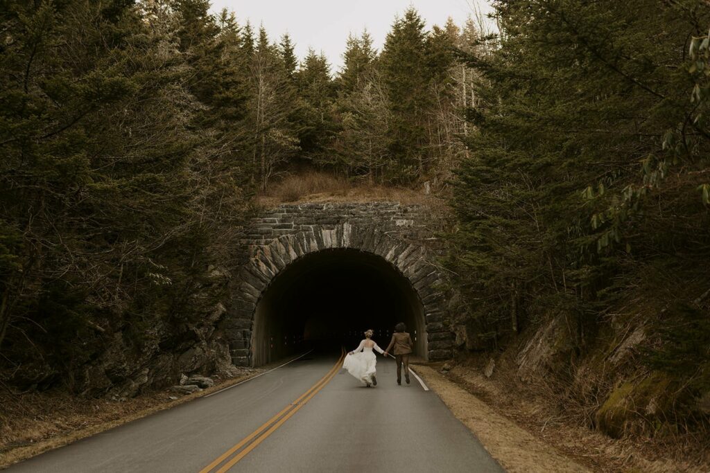 A couple is running towards a tunnel on the road during their Blue Ridge Parkway Wedding in their nice outfits with pines all around them.