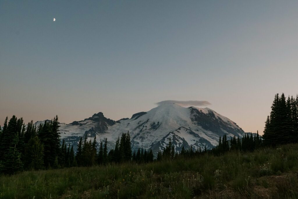 A night time view of Mount Rainier in Washington State with the moon and sunset behind the mountain.