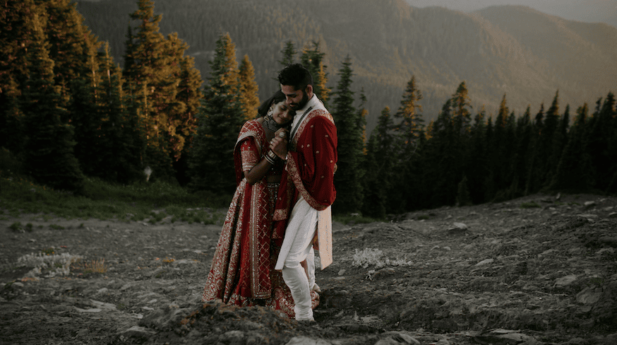 A couple stands close holding hands on a steep rocky mountain with evergreens in the background during their elopement. They are wearing bright red colors in traditional Indian clothing and are in Washington for their wedding.
