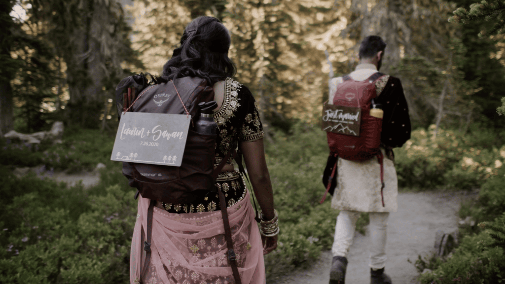 A couple hikes through a wooded trail during their elopement with backpacks on, traditional Indian clothing, and signs on their back saying “just married” and their names.