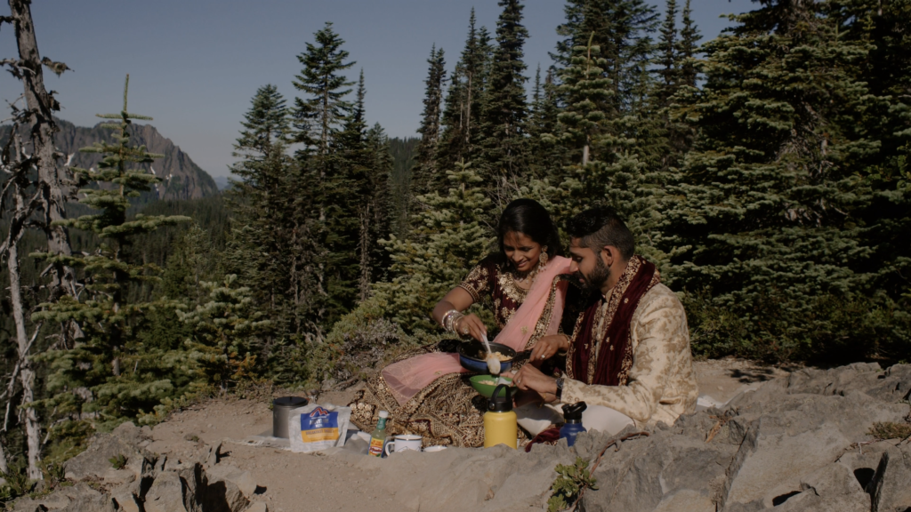 A couple sits having a picnic during their national park wedding in the morning. They are making backpacking eggs on a rocky area surrounded by evergreens.