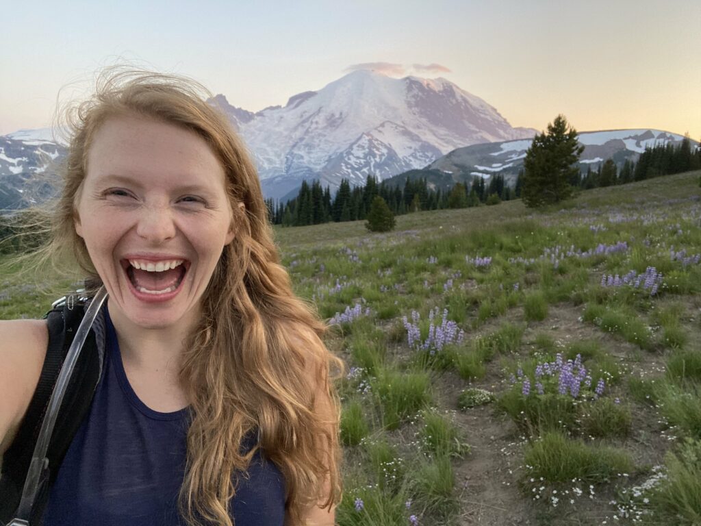 Kathryn, a photographer and videographer for Evergreen Era Films, stands in front of a sunset view of Mt Rainier national park while photographing an elopement. She has a big smile on her face and a backpack on. The mountain is glowing purple. Kathryn specializes in Mount Rainier Elopements