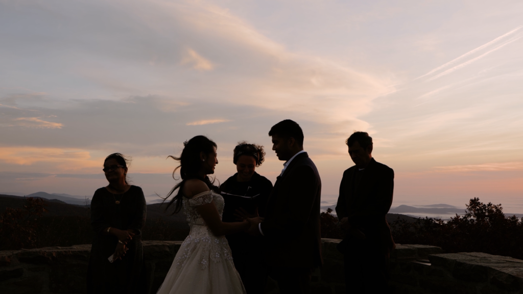 A couple, an officiant, and two witnesses are standing in front of a beautiful sunrise sky during a ceremony. They are silhouetted by the sky.