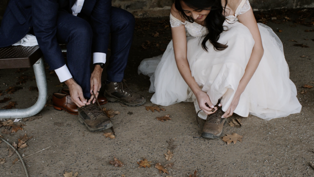 A couple with wedding clothes on are putting on and lacing up some hiking boots for their adventure wedding in Shenandoah National Park.