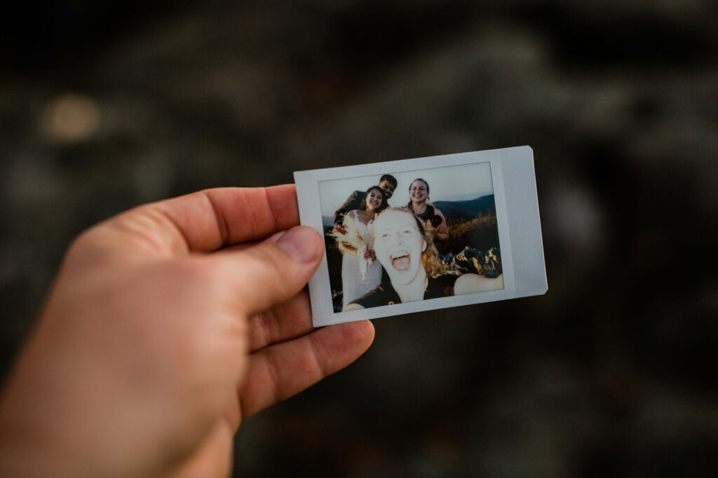 A hand is holding a polaroid image with a few people in the image. They are all smiling at the camera and one person is holding it and over exposed.