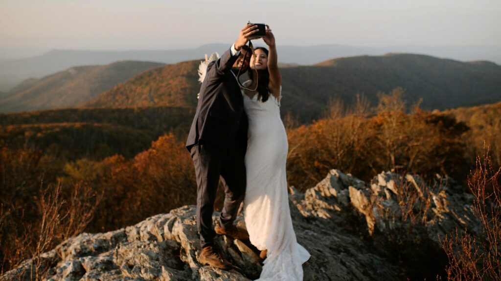 A couple is taking a selfie in front of an orange mountain view with their instant camera during sunset while having their wedding in Shenandoah National Park.