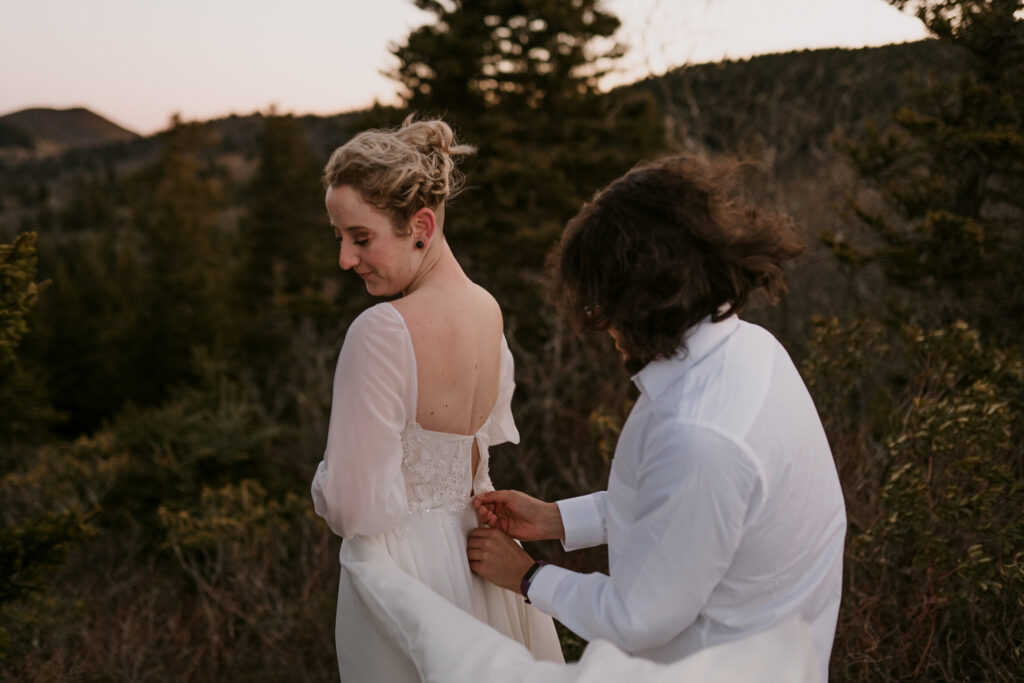 A man helps a woman get zipped into her wedding gown before their intimate sunrise elopement on a mountain.