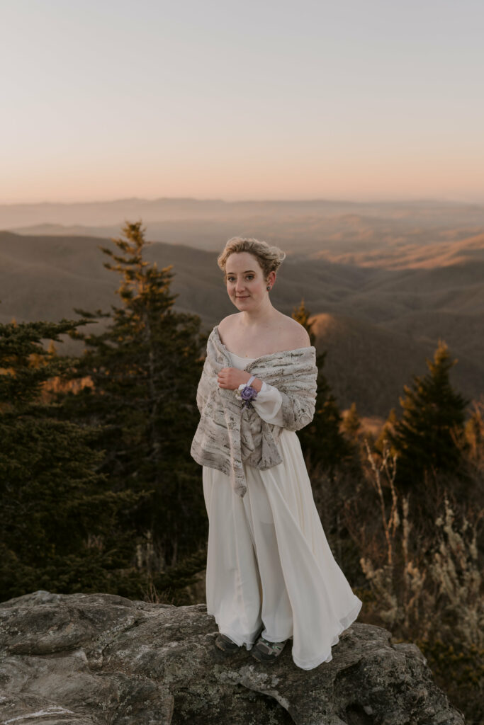 A bride in a wedding gown smiles on top of a rock at her intimate sunrise wedding in the mountains.