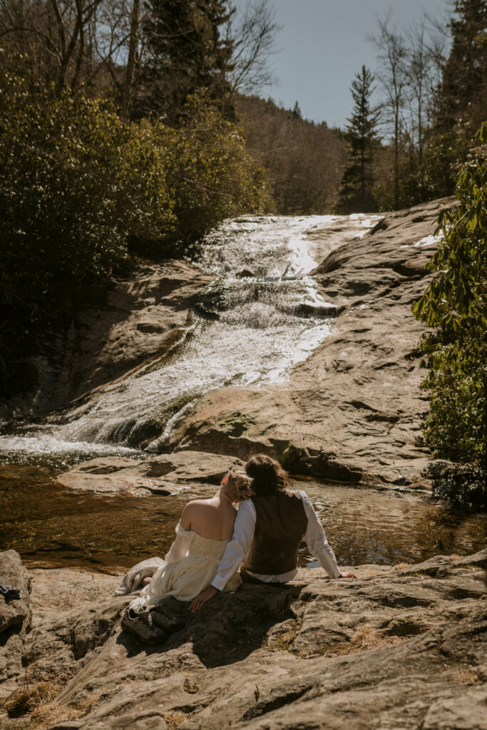 A woman rests her head on her husband’s shoulder while sitting on a rock in front of a waterfall in the sun.