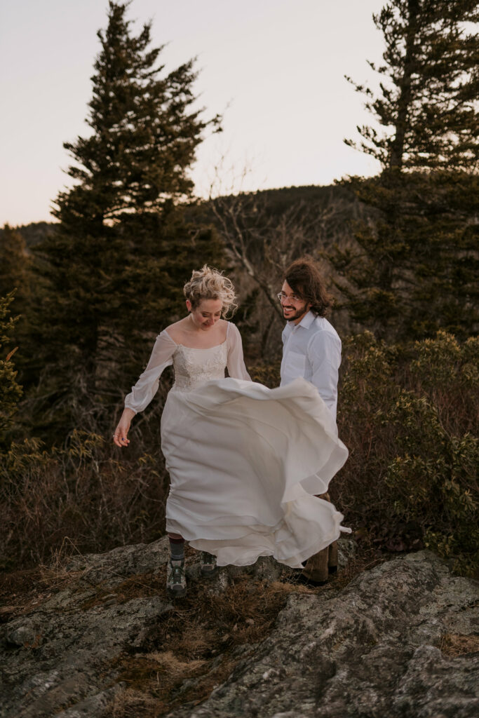 A man and a woman in wedding clothes stand on a mountain while the bride’s dress blows in the breeze as they get ready for their intimate sunrise wedding.