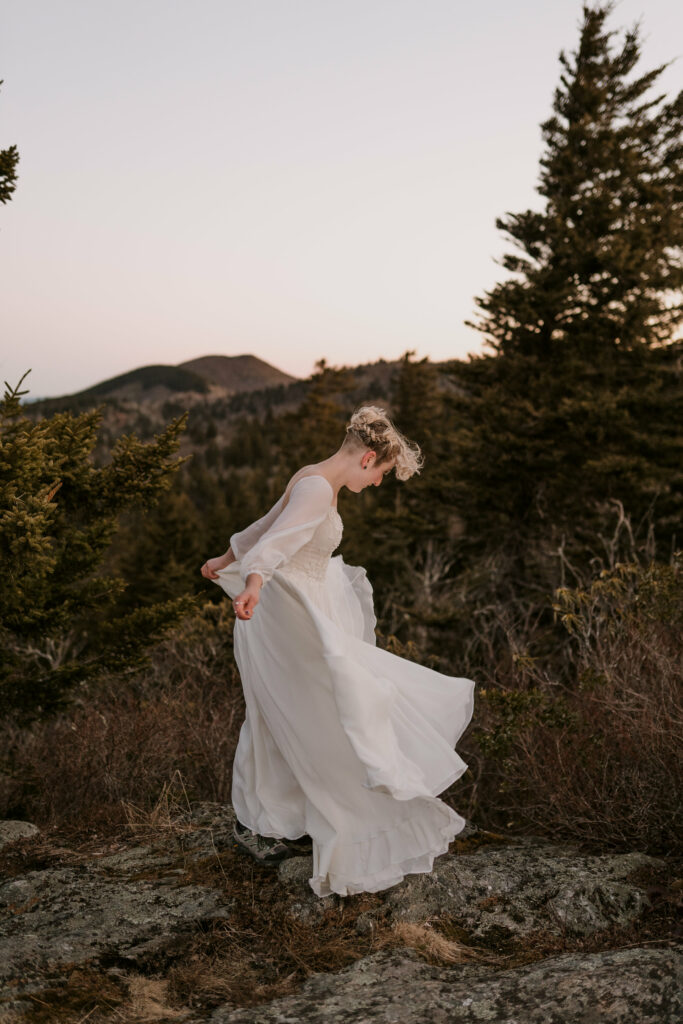 A woman' stands on a mountain wearing a white wedding gown that blows in the breeze during her intimate sunrise wedding.