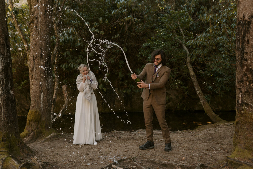 A man pops a champagne bottle in the woods while his wife stands by and laughs.