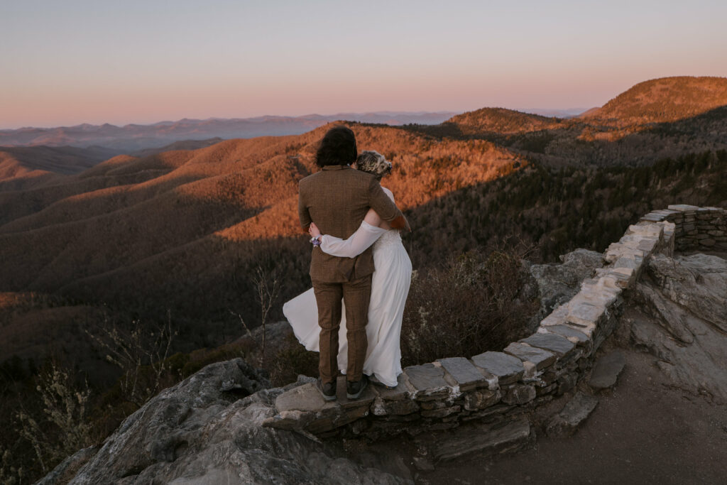 A couple stands on the ledge of an overlook with mountains in front of them during their Intimate Sunrise Wedding