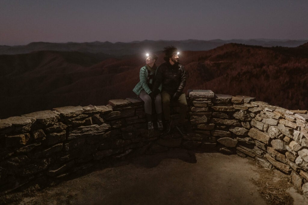 A couple sits together before their elopement in the early morning hours wearing headlamps with a mountain range in the background before their intimate sunrise wedding.