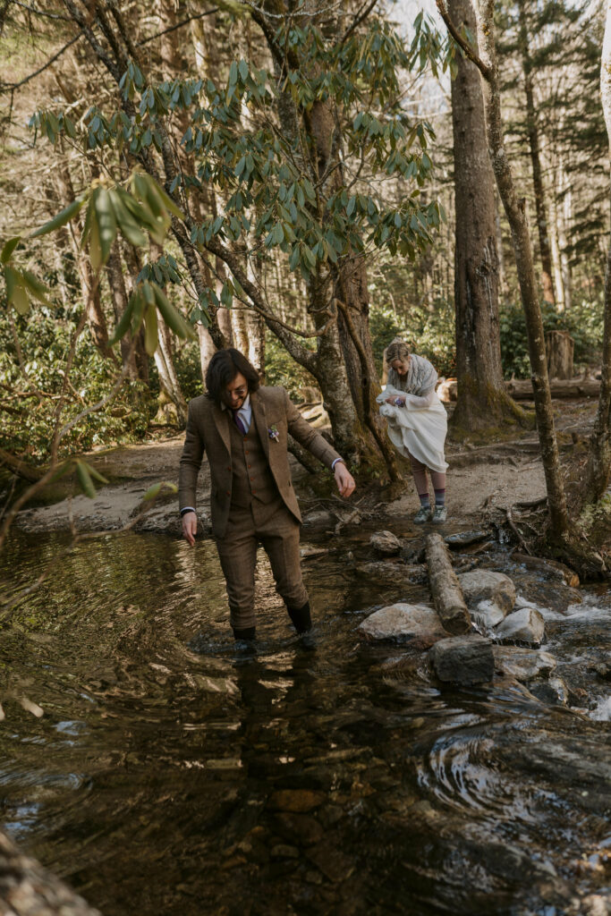 A man and woman in wedding clothes walk across a creek in the woods.