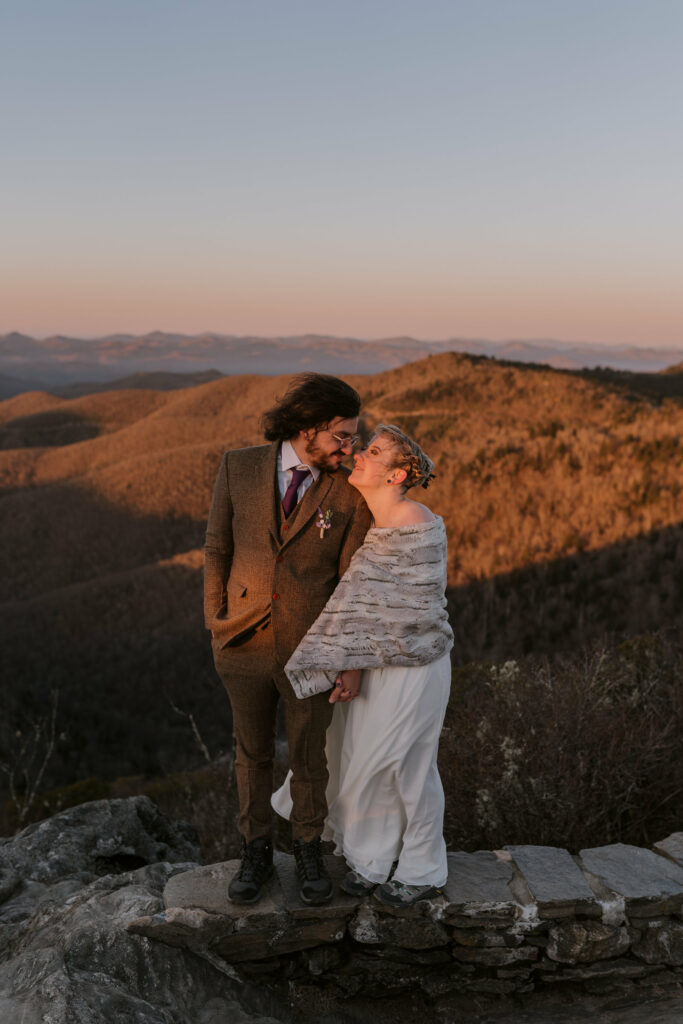 A man and woman hold their heads close together as they cuddle on top of a mountaintop in wedding clothes for their intimate sunrise wedding.
