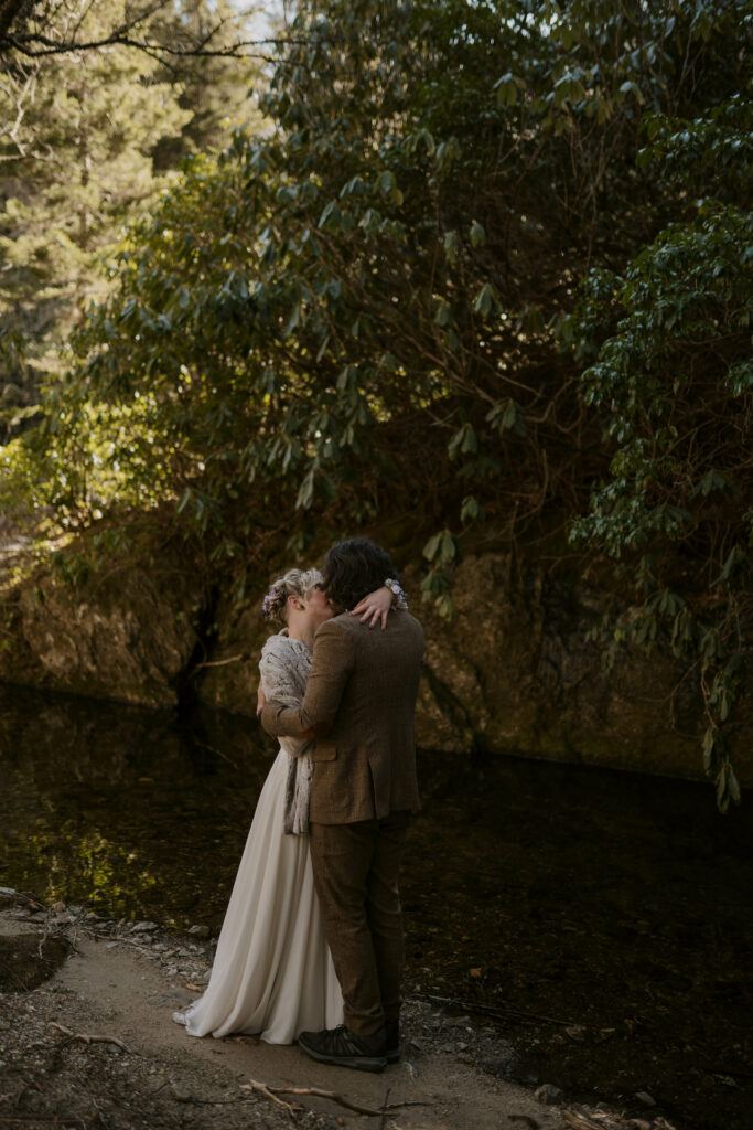 Husband and wife have their first kiss alongside a riverbank in the woods.