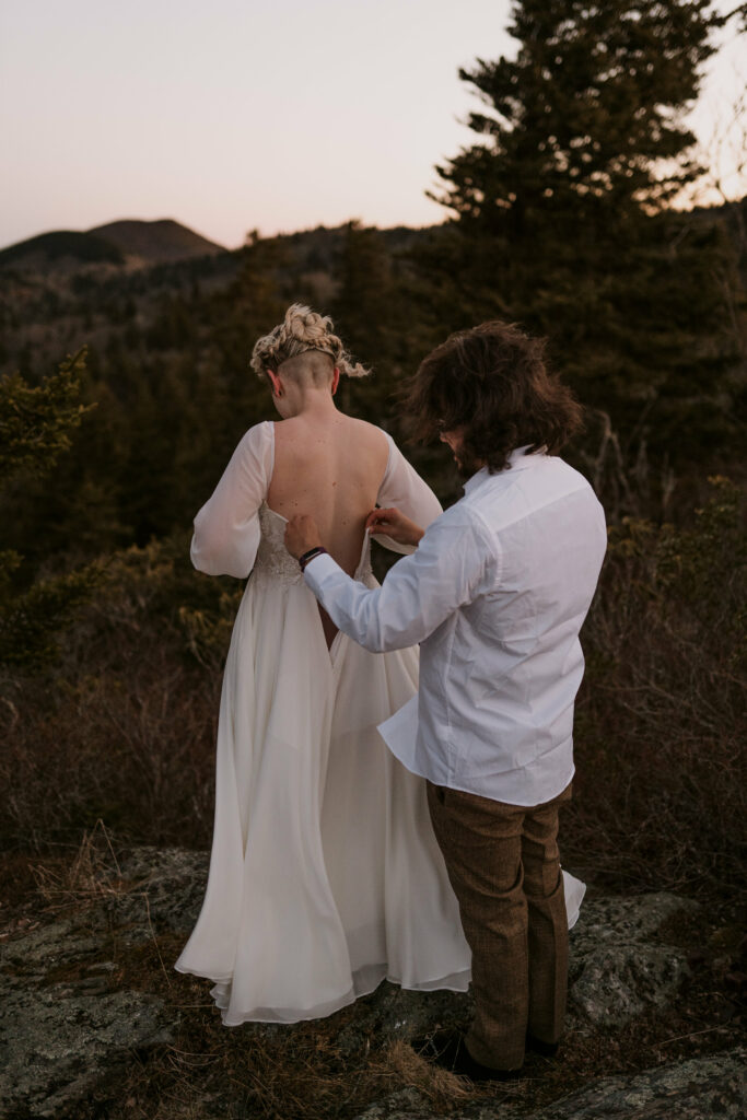 A man helps a woman into her wedding gown while standing on a mountain before their intimate sunrise wedding.