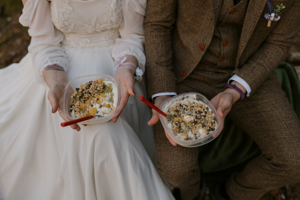 A couple in their wedding outfits holding bowls of granola and yogurt during their intimate sunrise wedding.