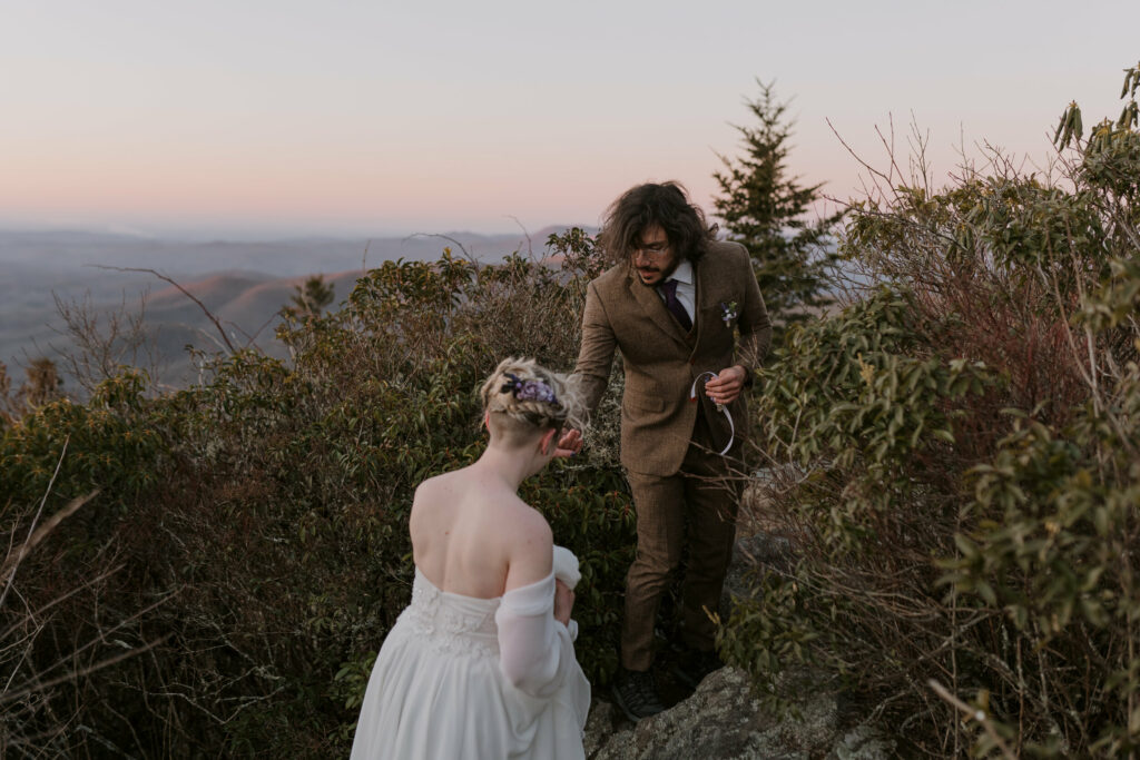 A man helps a woman walk up the side of a mountain during intimate sunrise wedding.