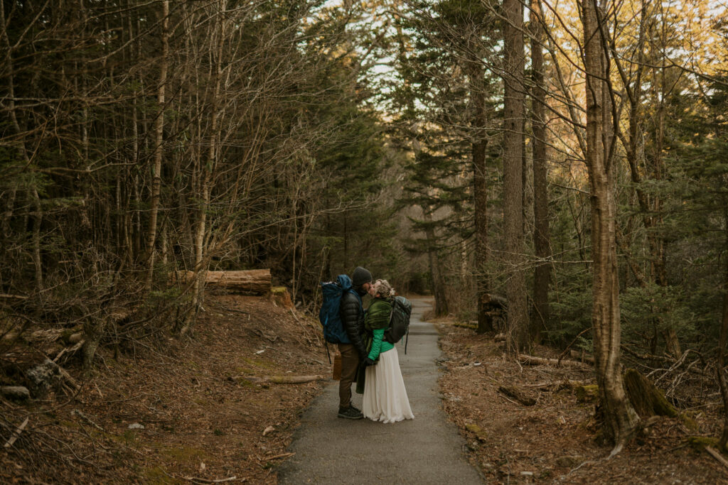 A man and wife in wedding clothes and hiking gear stops on a trail in the forest to kiss during their intimate sunrise wedding.