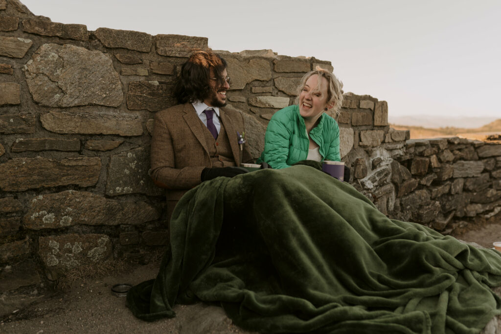 A couple sits under a green blanket on the edge of a mountain overlook and laughs during their intimate sunrise wedding.