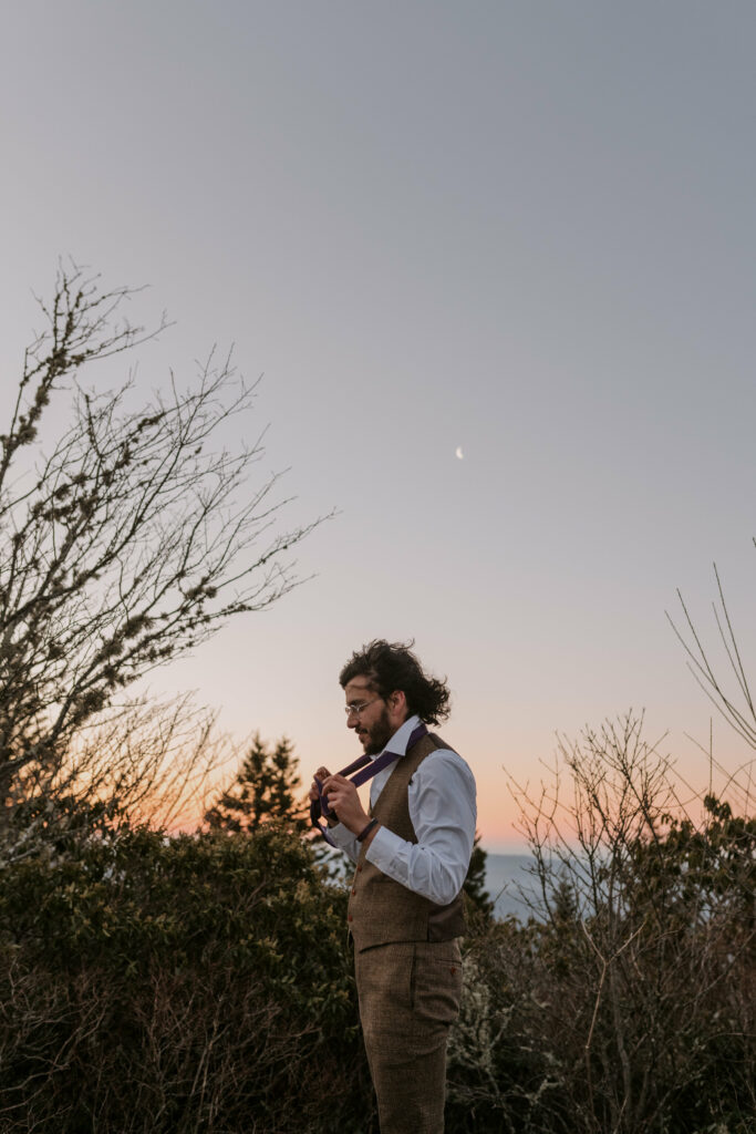 A man stands in front of the mountains with the sun coming up behind him while he ties his tie on his intimate sunrise wedding day.