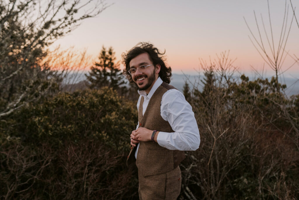 A man buttons up the vest with trees and mountains in the background before his intimate sunrise wedding.