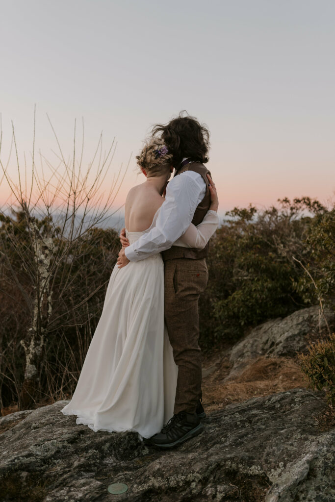Husband and wife in wedding clothes hugging on top of mountain at intimate sunrise wedding.
