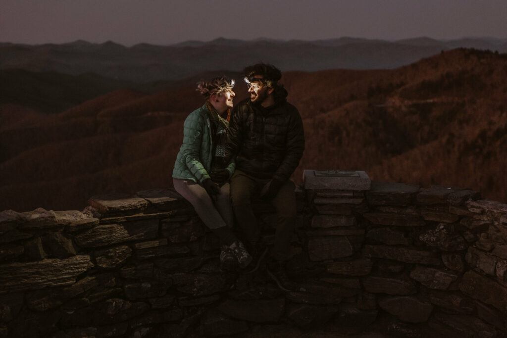 A couple sits on a mountaintop wearing headlamps and smiling at each other as the sun starts to rise over the mountains in the background during their intimate sunrise wedding.