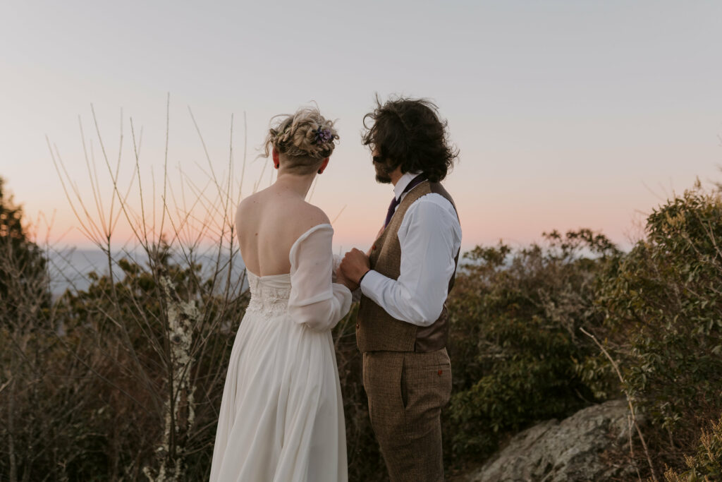 A man and woman hold hands and overlook a mountain range before their intimate sunrise wedding.