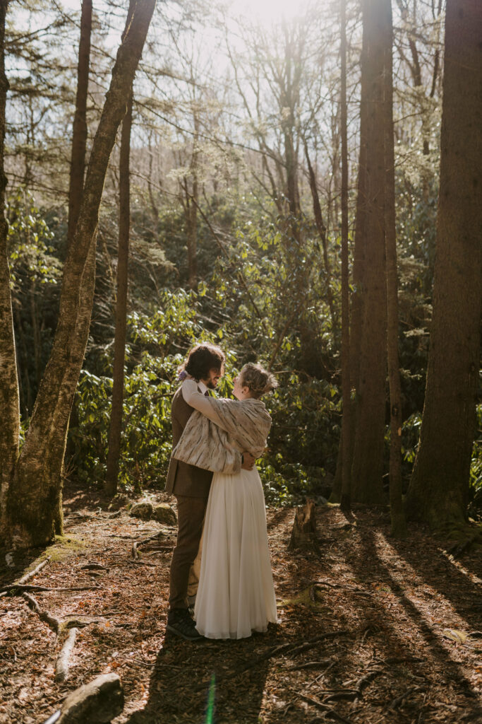 A newlywed couple hugging in the forest with pine trees and sun behind them during their intimate sunrise wedding.