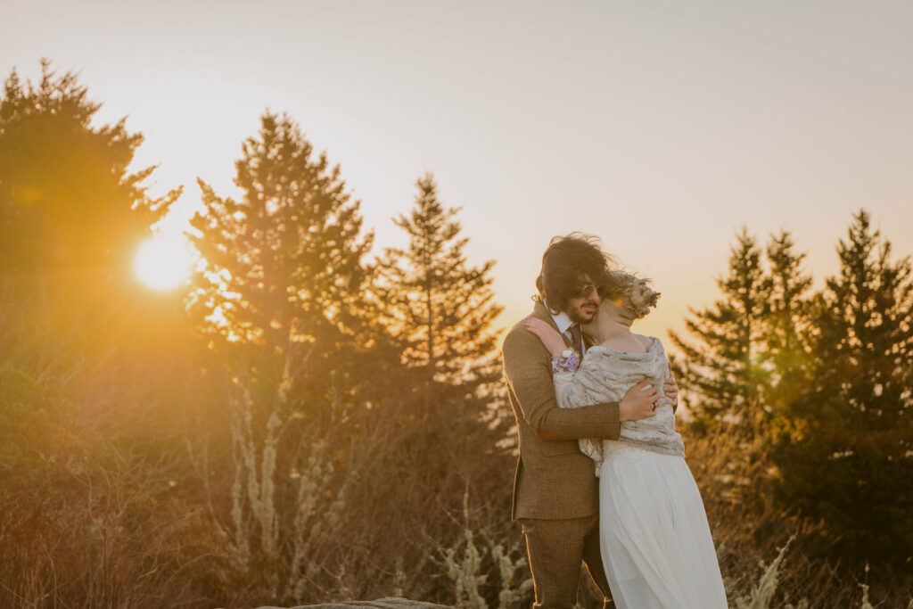 Husband and wife hugging with the sun shining through the pine trees behind them during their intimate sunrise wedding.