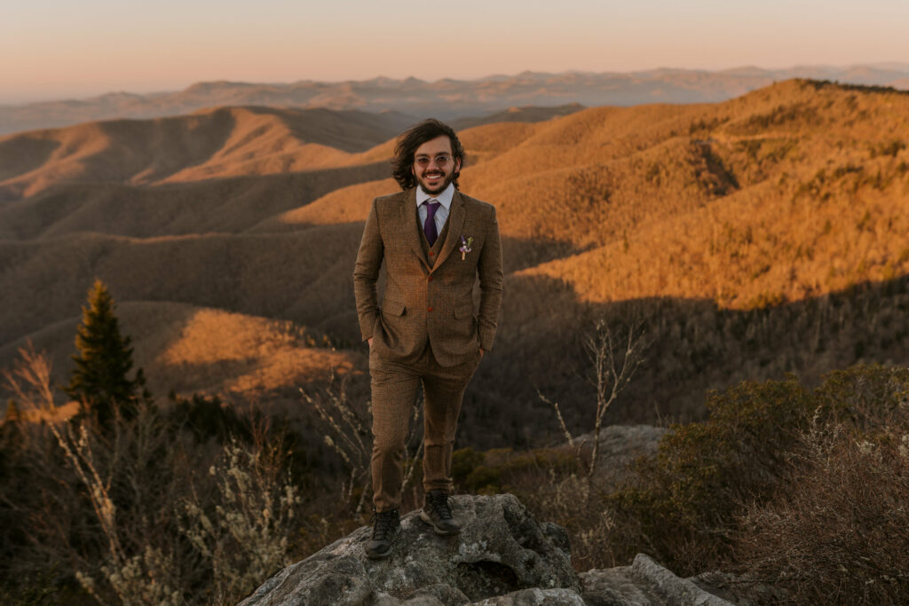 A man in a suit and sunglasses smiles on mountain for a portrait at his intimate sunrise wedding.
