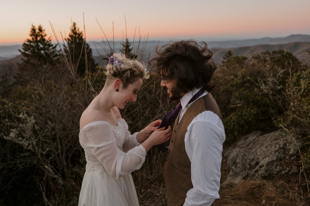 Woman helps man with his tie on top of a mountain at their intimate sunrise wedding.