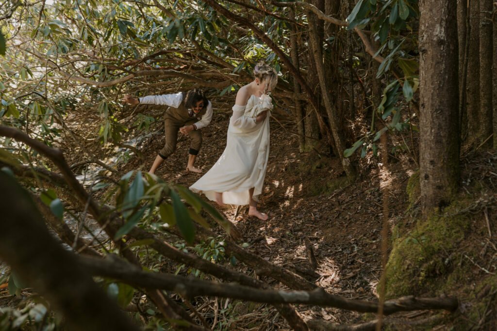 A couple walks through rhododendrons in the woods while wearing wedding clothes.
