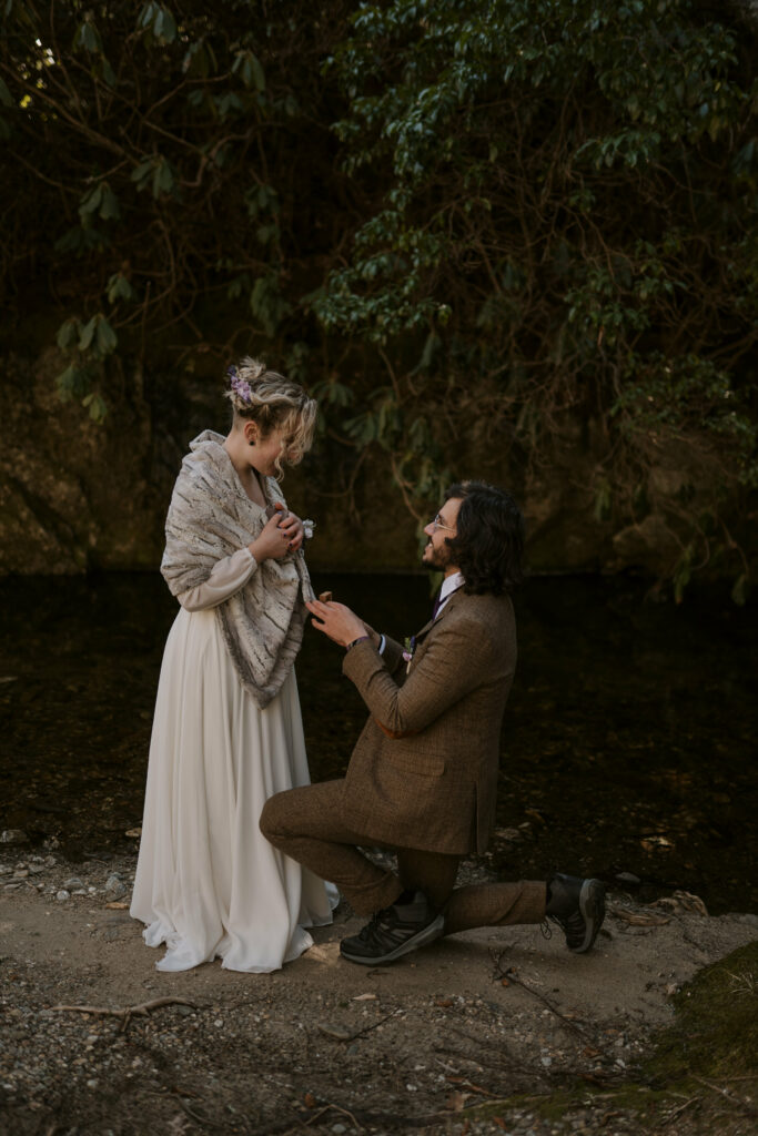 A man gets down on one knee in the woods in front of a creek as he marries his wife.