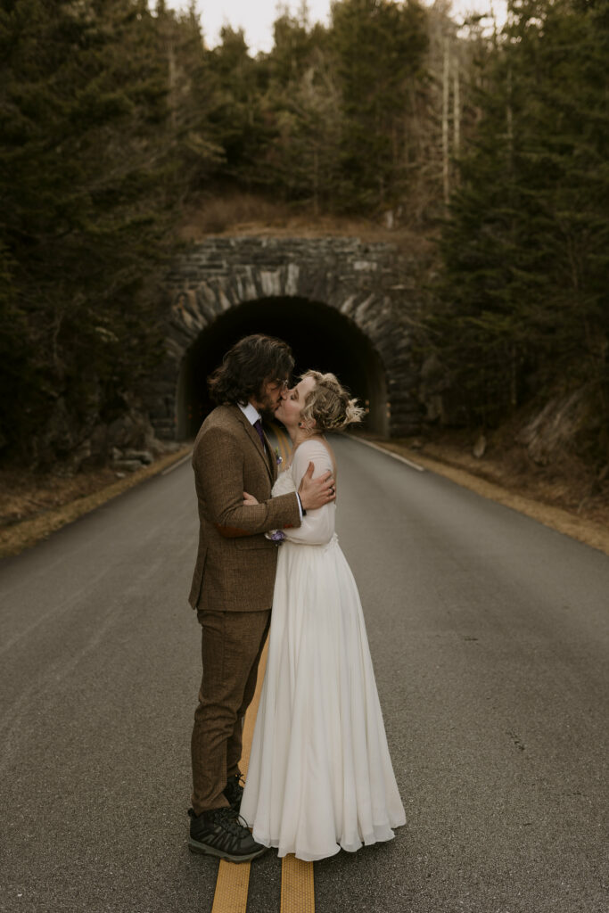 A newlywed couple kisses in the middle of the road in front of a tunnel in the forest during their intimate sunrise wedding.
