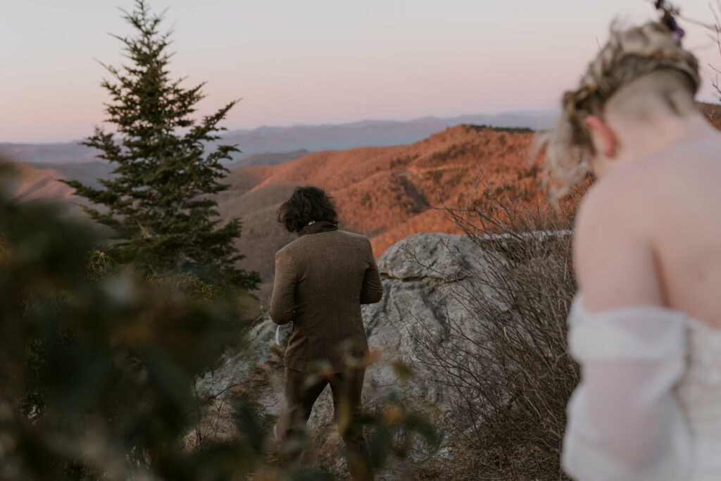 A couple is walking along a big rock at sunrise on the blue ridge parkway with pink lighting up the mountains for their wedding morning.