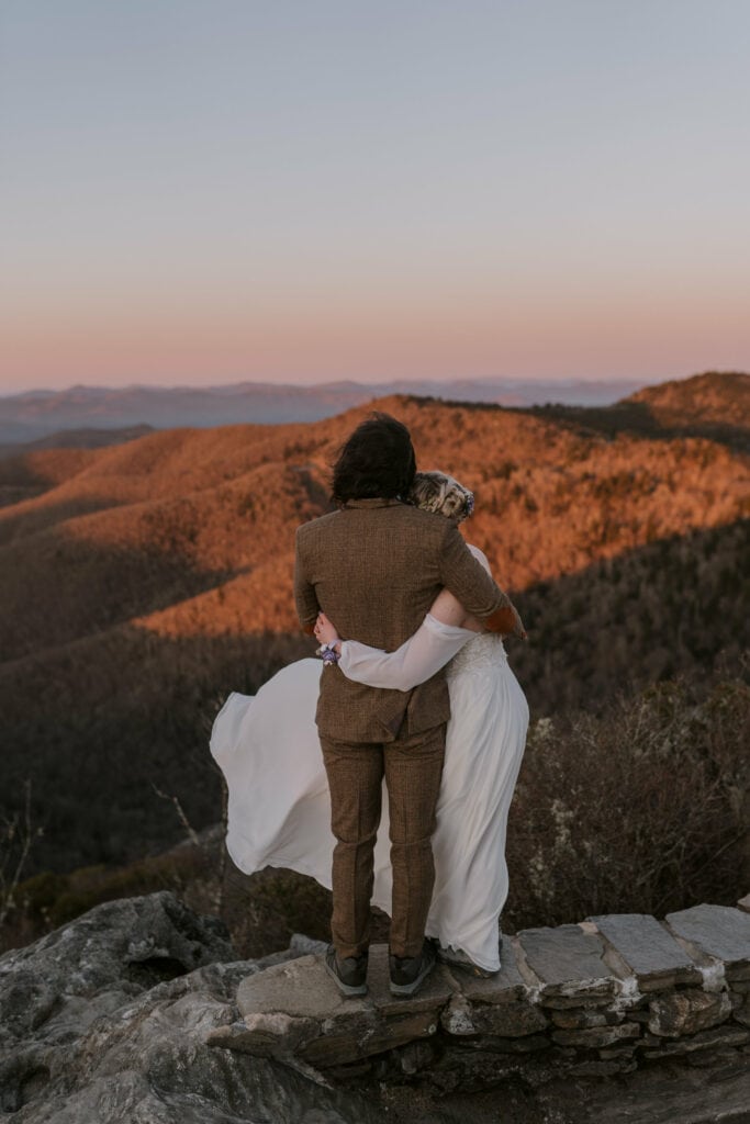 A couple hugs on top of a rock facing the sun coming up on the mountains in front of them during their intimate sunrise wedding.