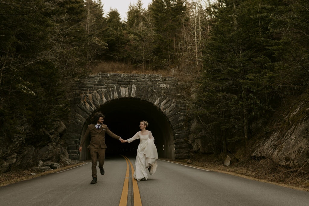 A newlywed couple holds hands and walks down the middle of the road in front of a tunnel in the mountains during their intimate sunrise wedding.