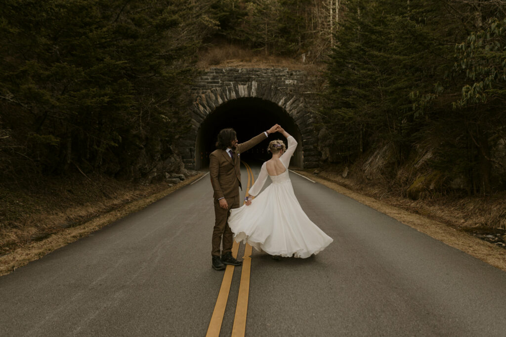A couple is dancing on the Blue Ridge Parkway, one of the best places to elope in asheville nc. They are getting married and the bride is twirling under the grooms arm. She is in a white dress and he is in a brown suit.
